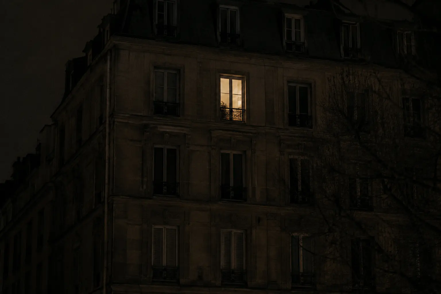 A single lit window on the upper floor of a dark stone apartment building at night